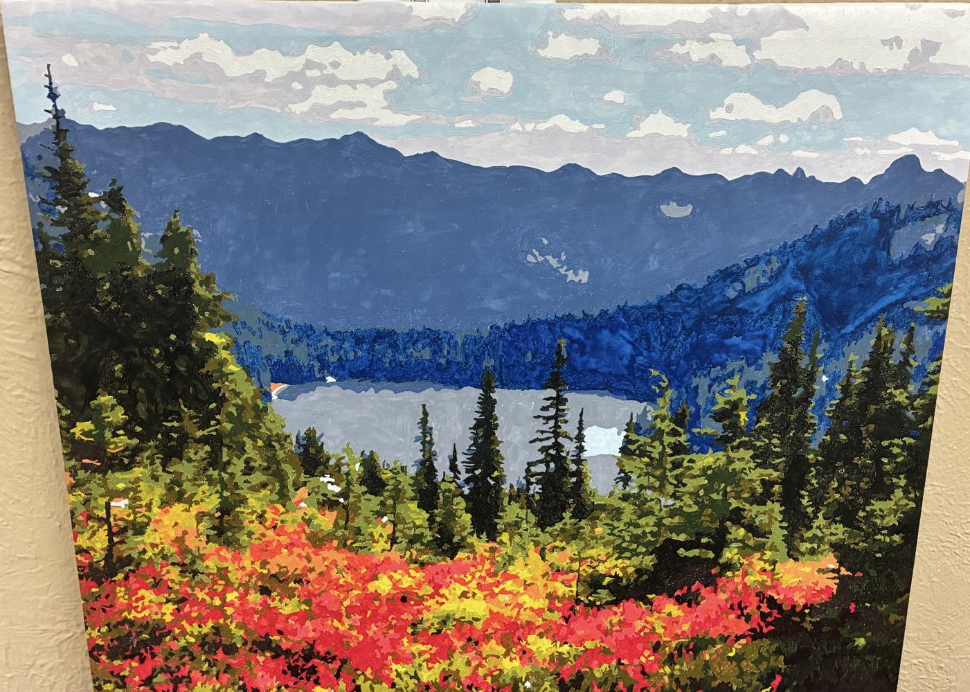 This artwork captures a breathtaking mountain landscape at the height of autumn. A tranquil lake rests between rugged peaks, reflecting the cool blues and soft grays of the distant mountains. The foreground bursts with vibrant fall color — deep reds, warm oranges, and golden yellows — framed by evergreen pines that add contrast and depth. Soft clouds drift across a blue sky, completing the peaceful, immersive atmosphere. This piece brings the beauty of untouched nature into any space. Its rich colors and serene composition make it ideal for living rooms, offices, cabins, or anyone who loves the outdoors.